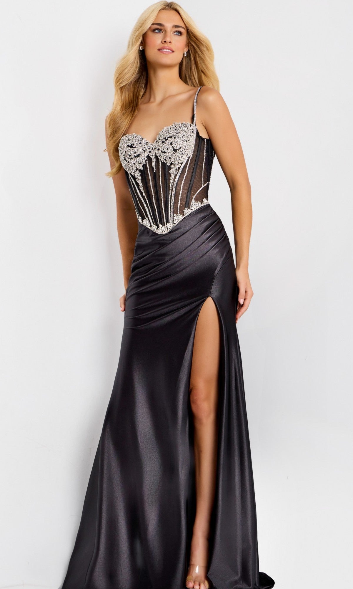 A woman in the Long Prom Dress: Jovani 47849, a black satin gown with a high slit and beaded corset bodice, stands against a plain white background.